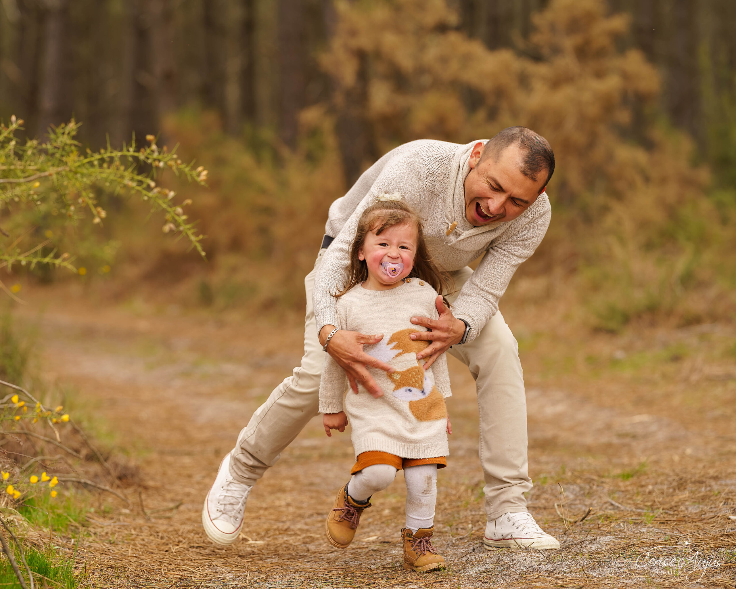 Père courant dans les bois pour attraper sa petite fille qui rit avec sa tétine lors d’une séance photo famille à Mimizan dans les Landes