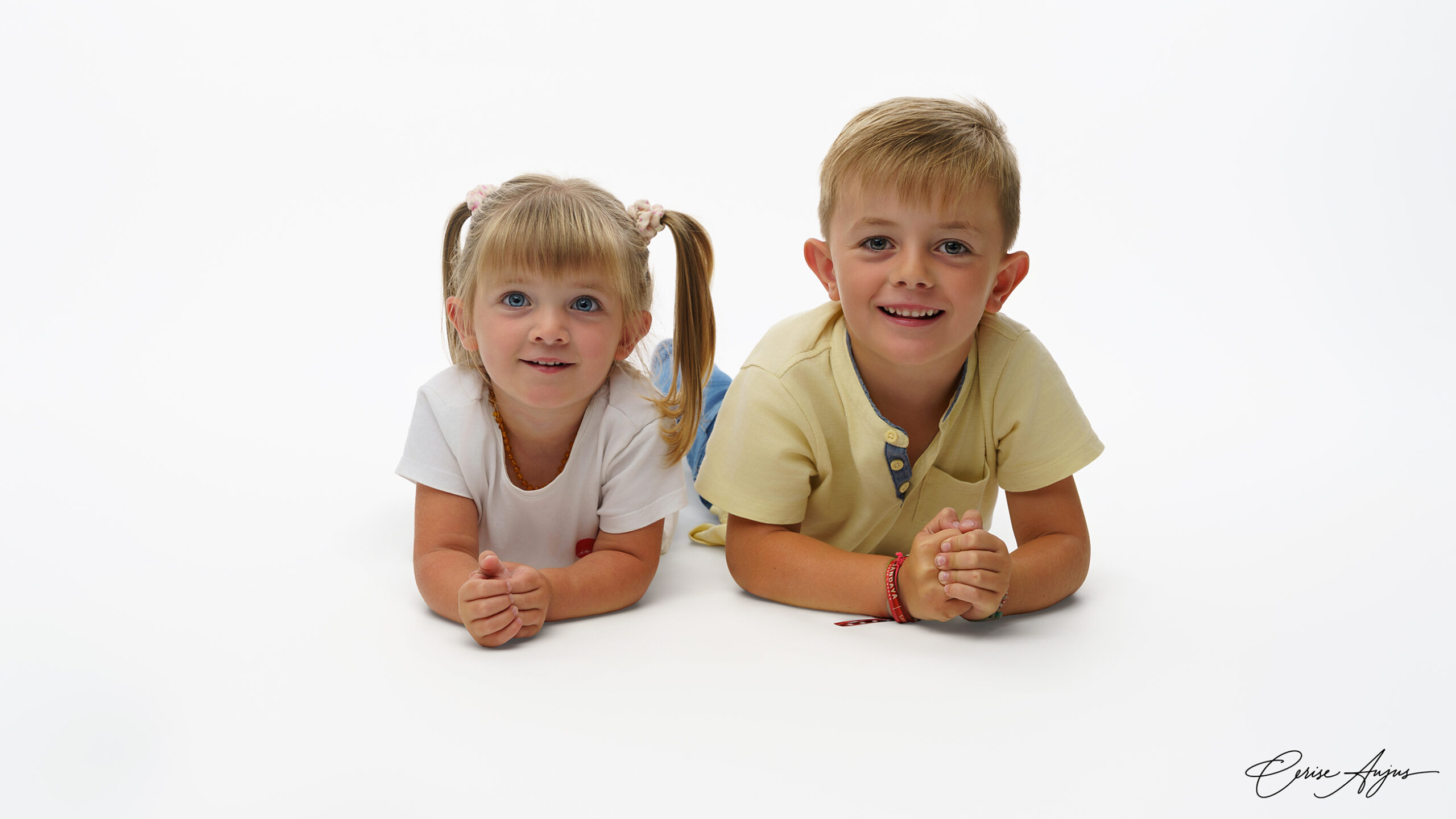 Deux enfants frère et sœur allongés sur le ventre sur fond blanc en studio, souriant face à l’objectif
