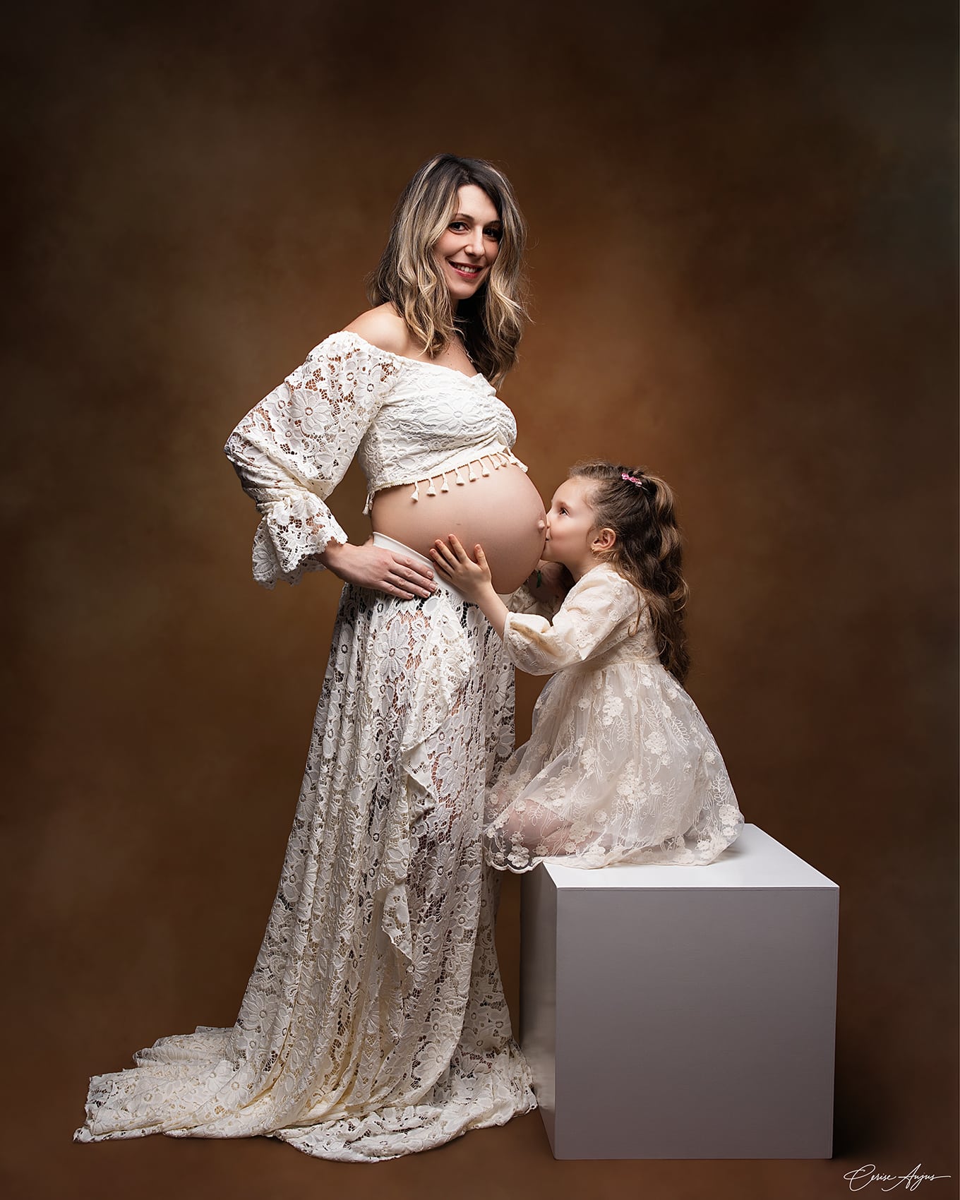 <br />
Fillette debout sur un cube de profil, mains posées sur le ventre de sa maman enceinte, faisant un bisou au ventre — séance grossesse avec enfant aîné dans les Landes