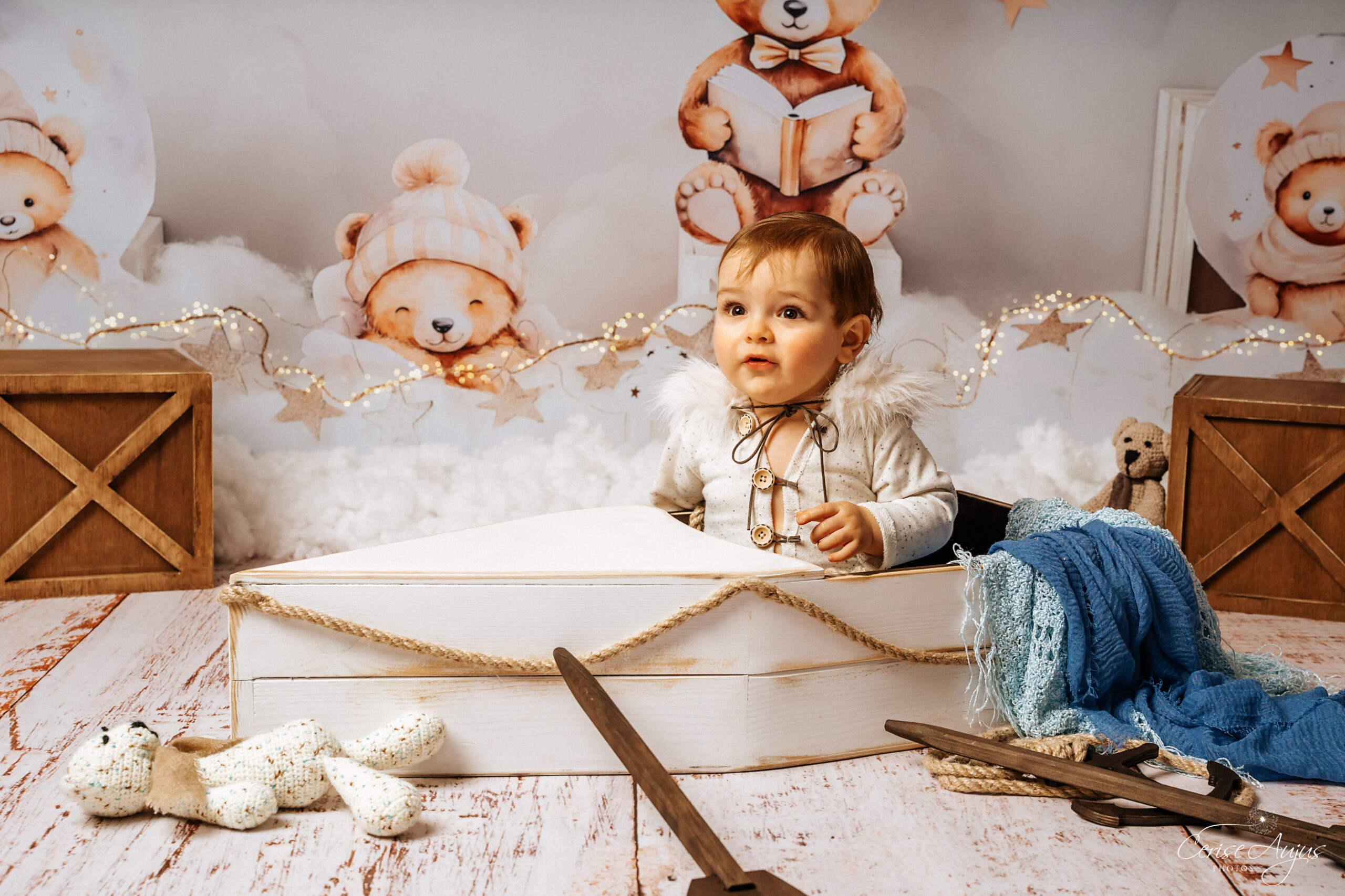 Petit garçon dans un bateau en bois dans un décor studio épuré avec nounours et nuages tons blancs crème et marron clair