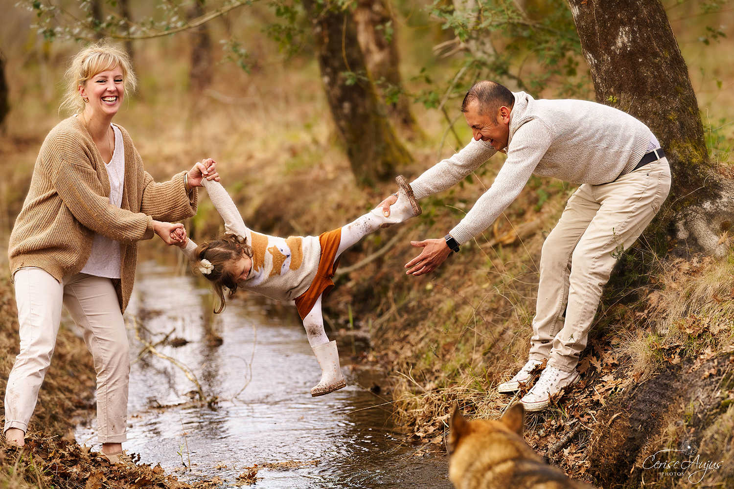 Père et mère tenant leur petite fille au-dessus d’un fossé rempli d’eau dans les bois à Mimizan, chacun par les mains et les pieds, lors d’une séance photo famille en extérieur