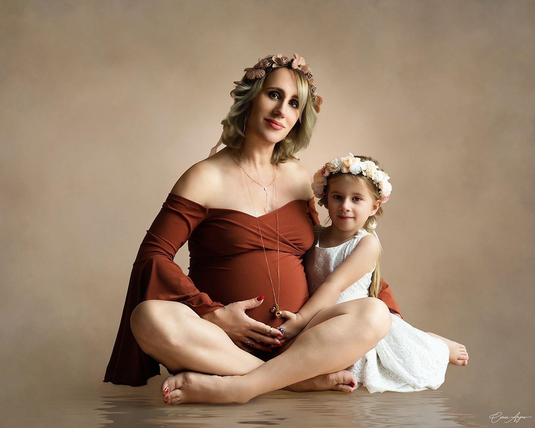 Fillette assise en tailleur au sol avec sa maman enceinte, main posée sur le ventre, regards tournés vers l'objectif — séance grossesse avec enfant aîné dans les Landes