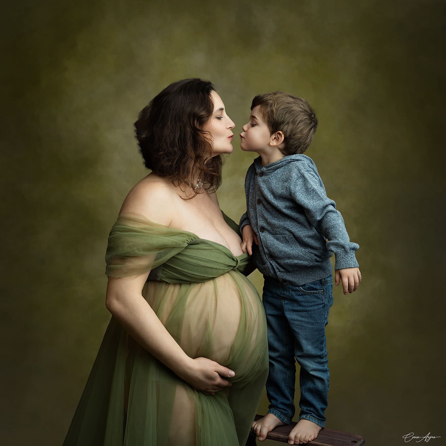 Femme enceinte en robe face à son fils debout sur un tabouret, leurs visages se rapprochent pour un bisou — séance grossesse avec enfant aîné dans les Landes