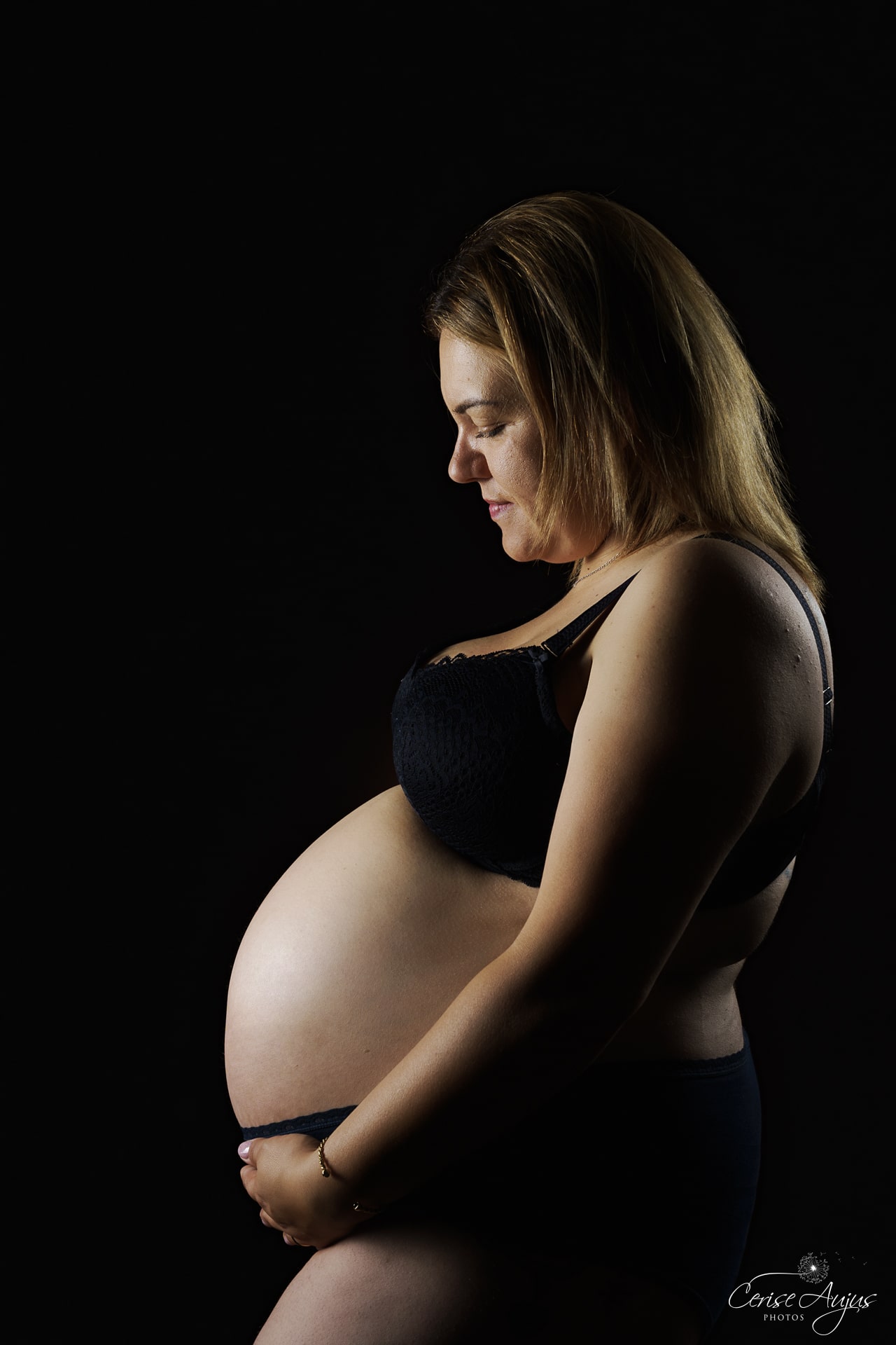 Femme enceinte de profil en lingerie noire sur fond noir, séance photo grossesse en studio près de Biscarrosse dans les Landes
