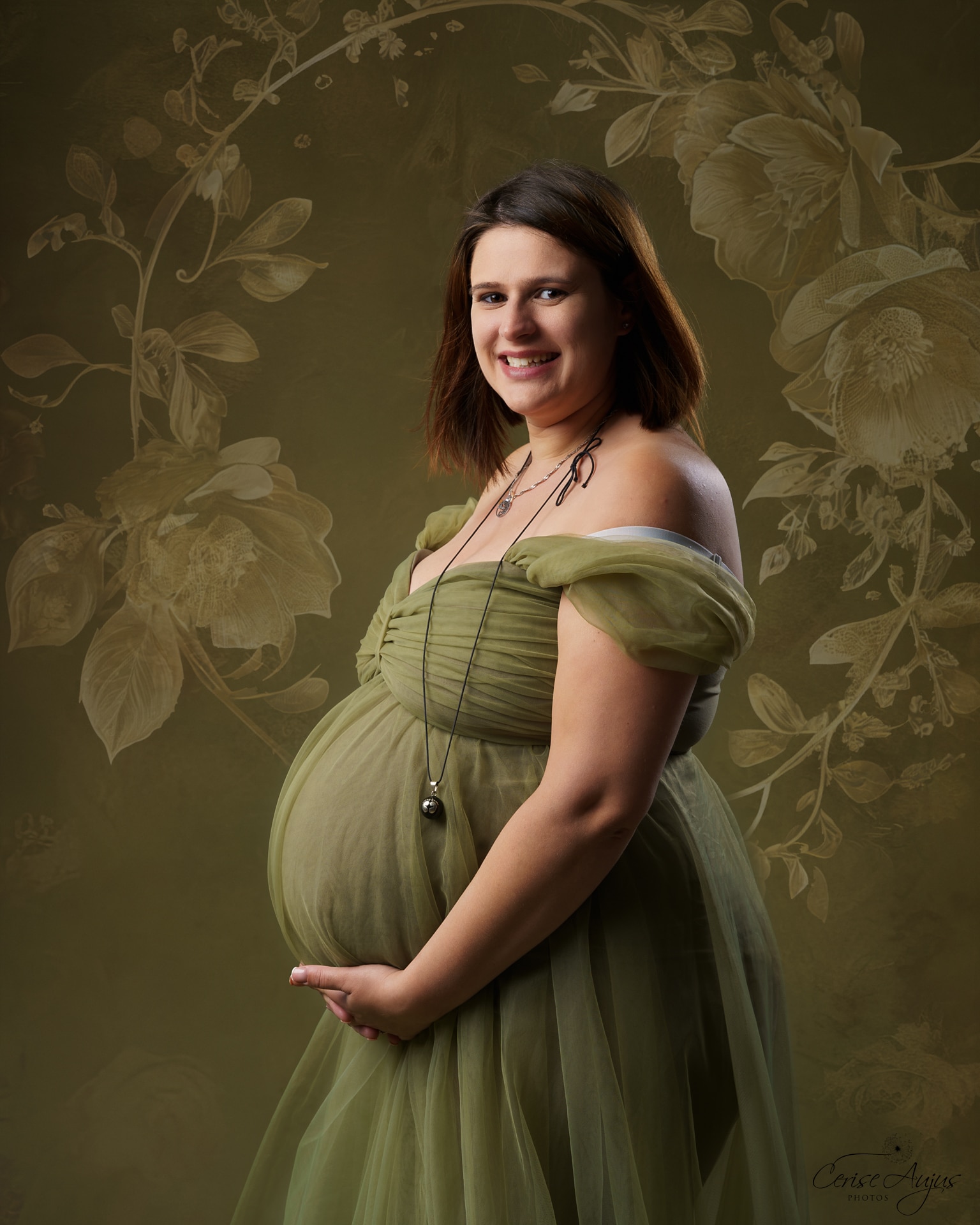 Femme enceinte souriante en robe verte sur fond fleuri kaki, séance photo grossesse en studio près de Biscarrosse dans les Landes