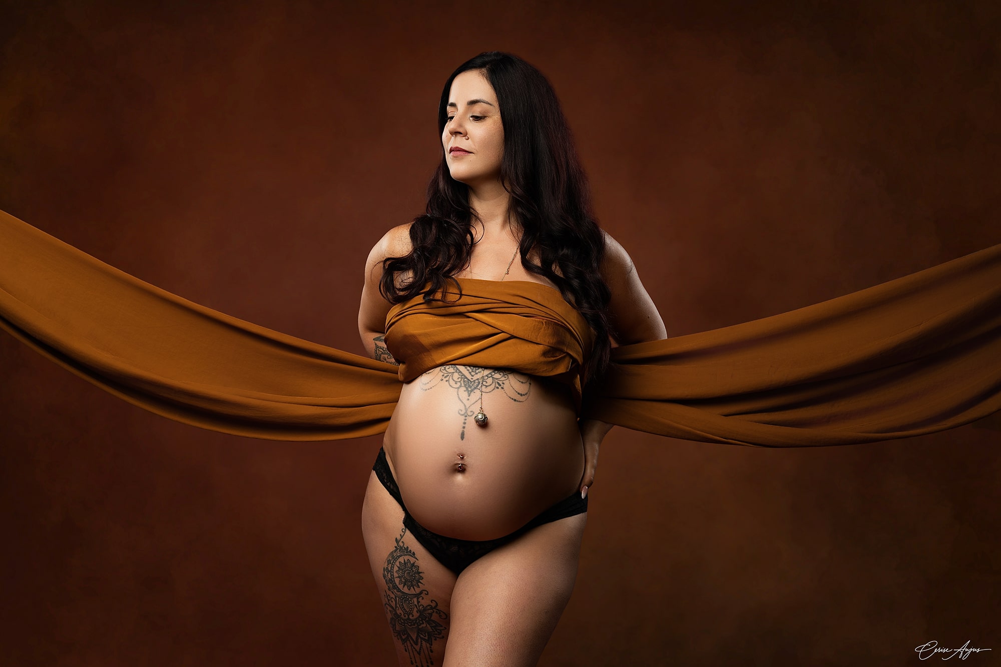 Femme enceinte en studio photo dans les Landes, voile cognac déployé de chaque côté, ventre rond orné d’un tatouage dentelle et d’un bola, fond brun chaud
